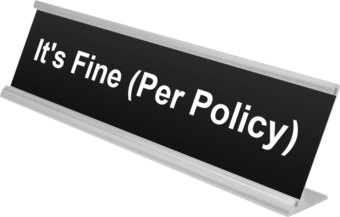 It's Fine (Per Policy) desk plate in black, shown in an aluminum desk holder