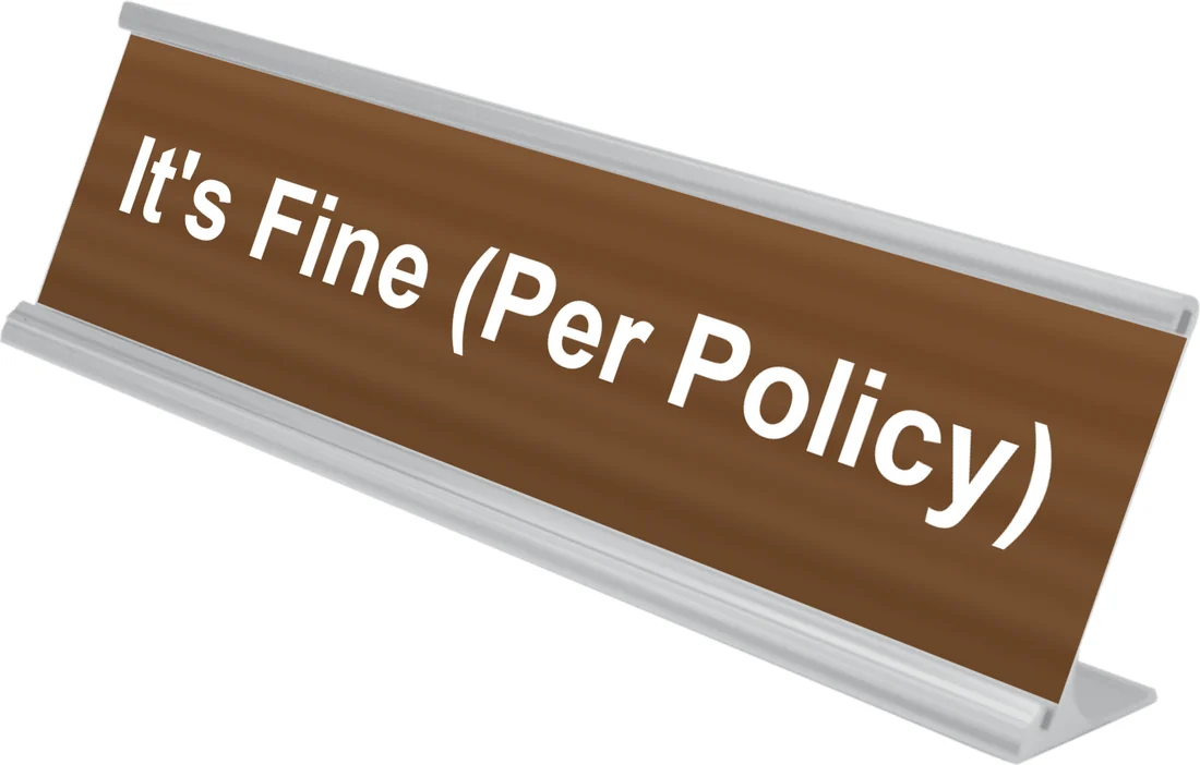 It's Fine (Per Policy) desk plate in walnut, shown in an aluminum desk holder