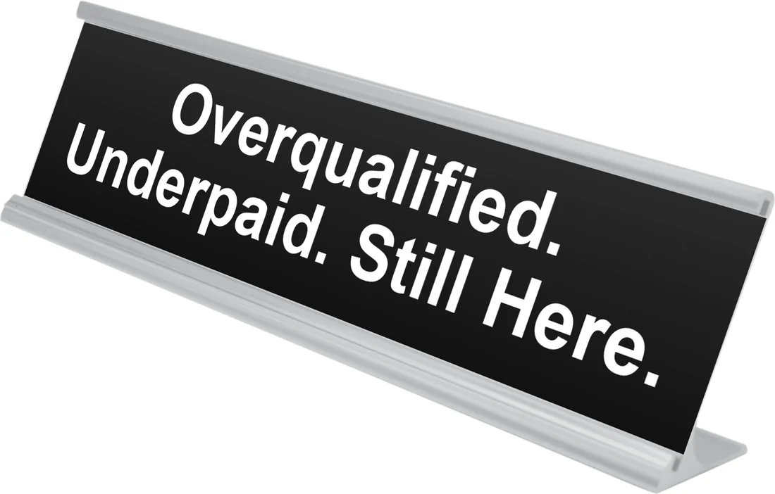 Overqualified. Underpaid. Still Here. desk plate in black, shown in an aluminum desk holder