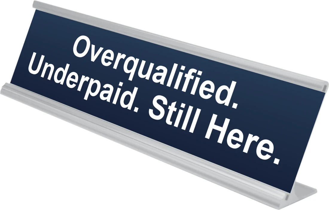Overqualified. Underpaid. Still Here. desk plate in navy, shown in an aluminum desk holder