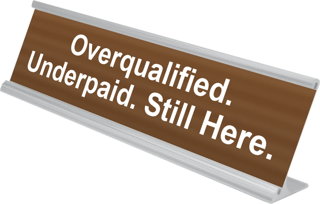 Overqualified. Underpaid. Still Here. desk plate in walnut, shown in an aluminum desk holder